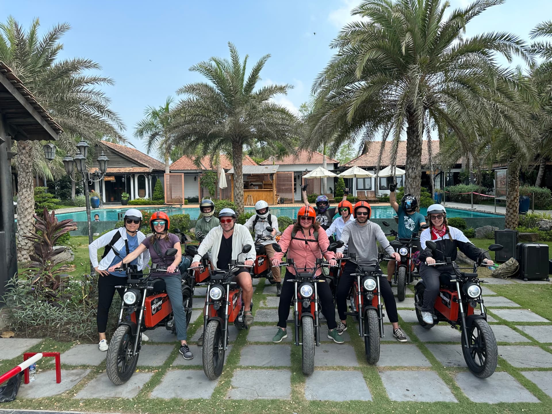 A smiling tour group in helmets posed on red electric motorbikes outside a tropical resort with palm trees and a pool