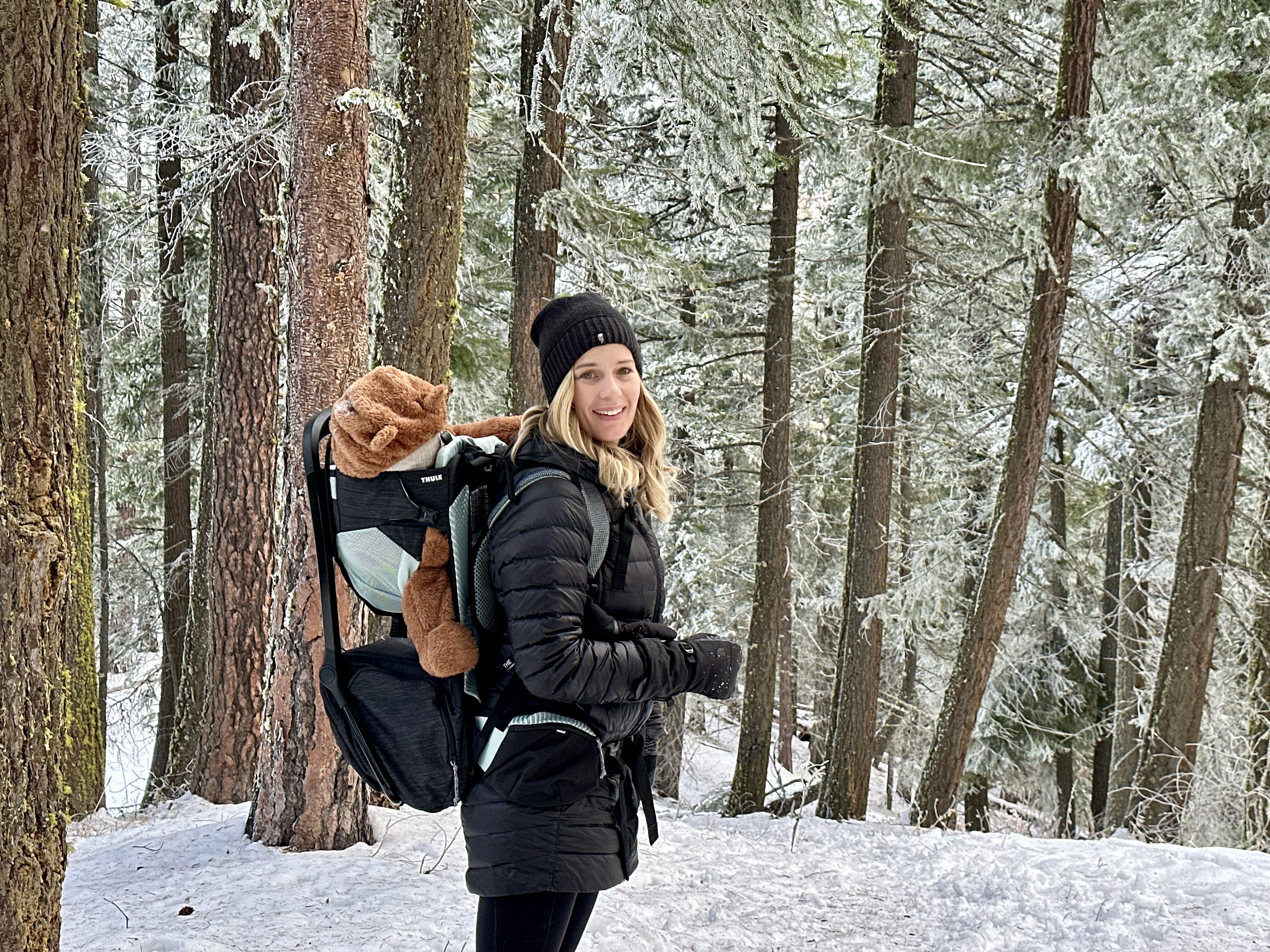 A smiling hiker carries a child in a backpack carrier through a snowy, frost-covered evergreen forest
