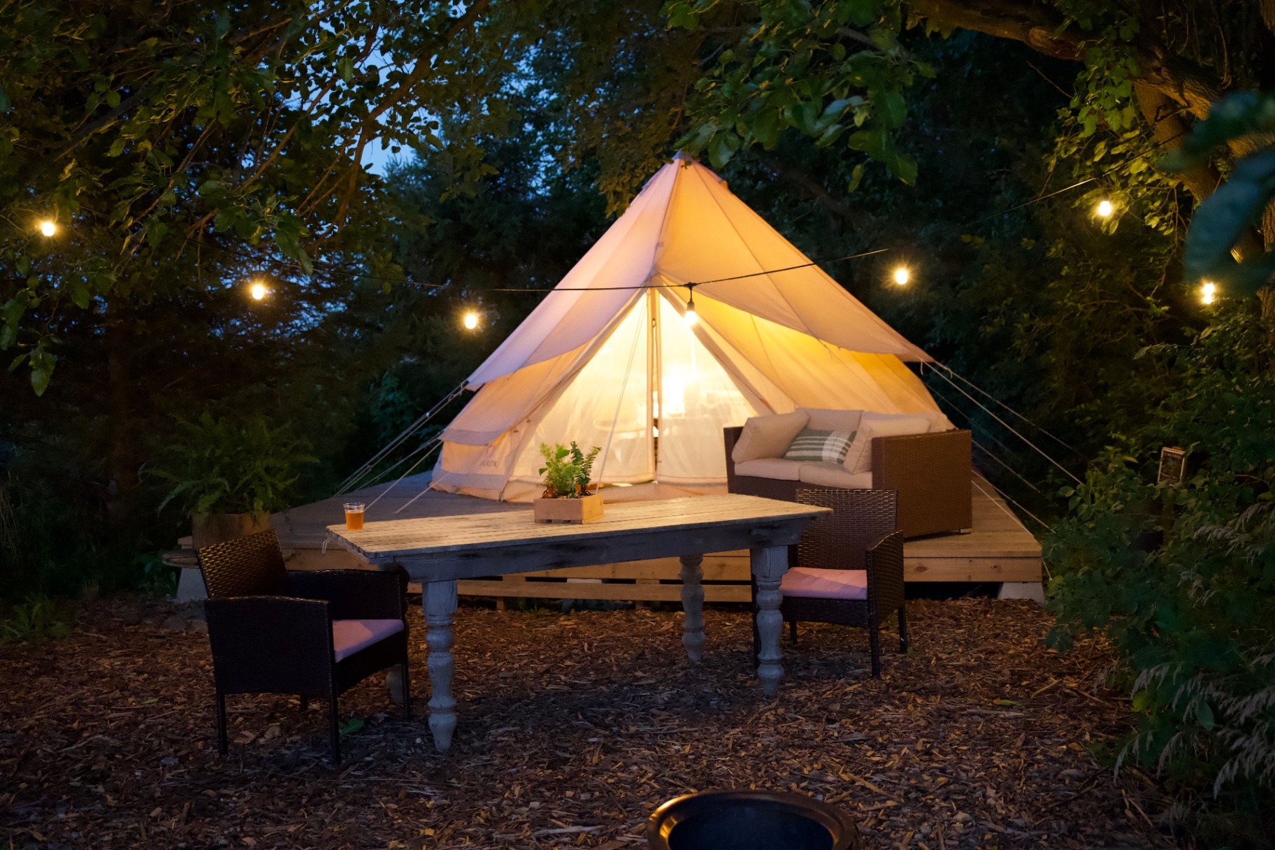 A glowing canvas glamping tent at dusk with string lights and an outdoor dining setup